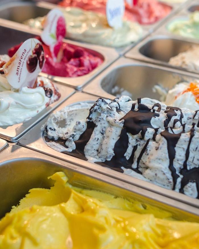 Eistheke mit vielen Sorten in Edelstahlbehältern, vorne Stracciatella mit Schokoladensoße